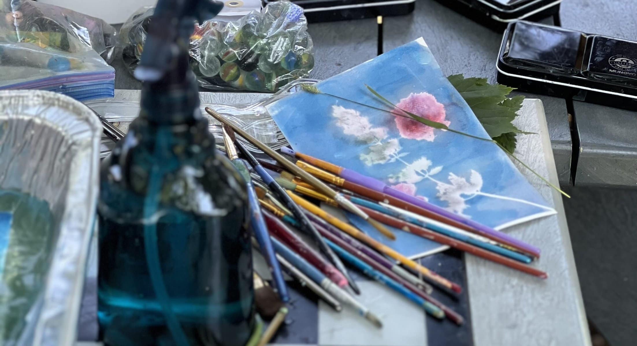 Close up of a partially painted cyanotype print surrounded by marbles, paint brushes, leaves, a spray bottle, and a foil pan