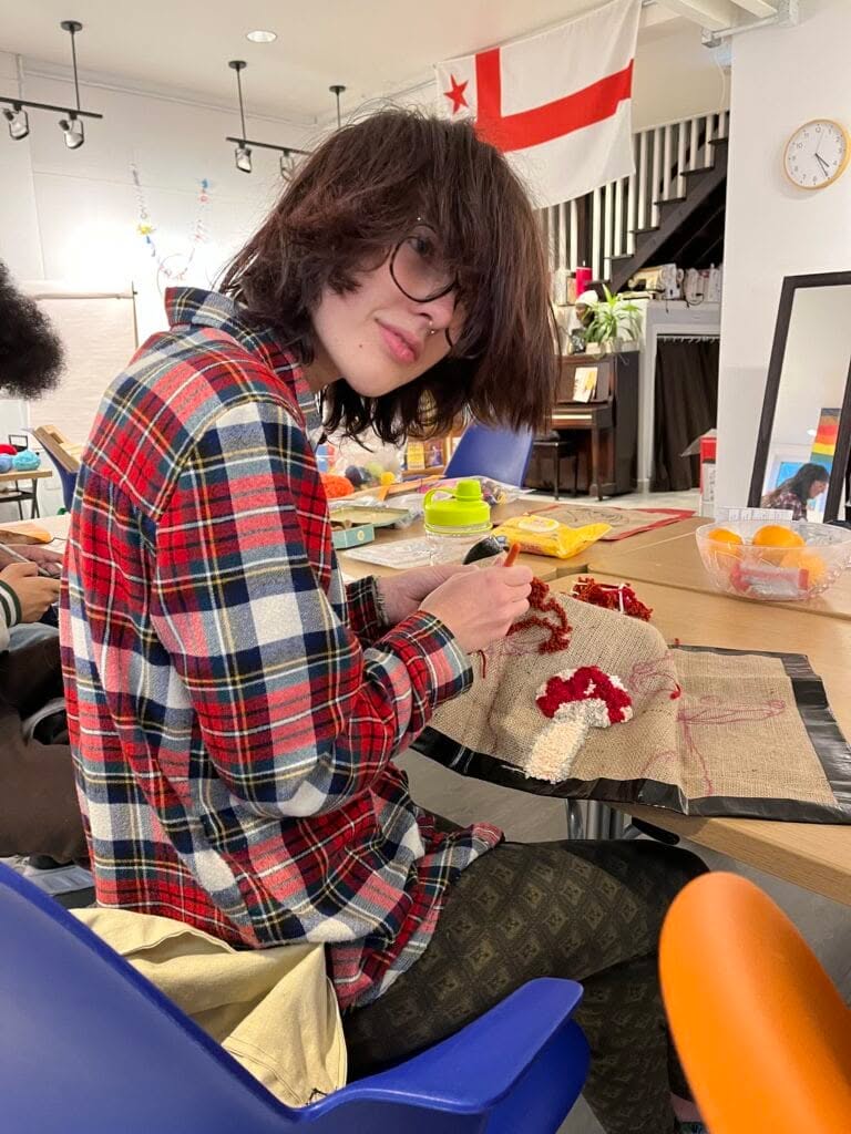 Photo of Caz looking through their long bangs at the camera while rug hooking a mushroom