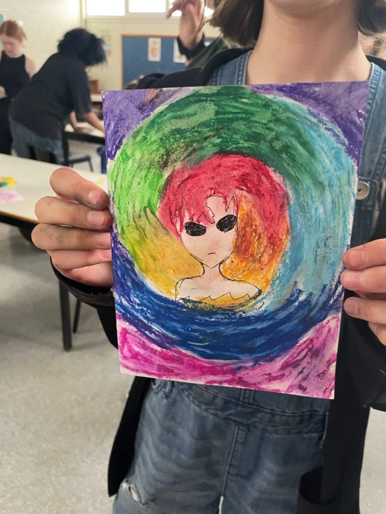 Youth holds up an illustration. There's a central figure with a rainbow swirl surrounding them. The swirl grows from their head like hair.